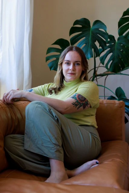 Portrait of a woman in a casual green top and wide-leg pants, relaxing on a brown couch with a monstera plant behind her.
