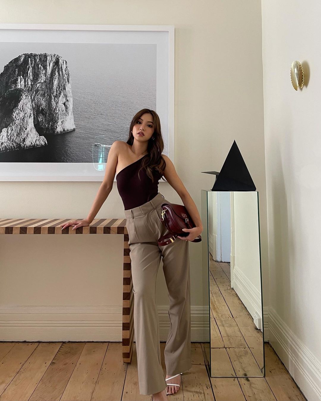 Woman poses indoors wearing a maroon one-shoulder top and beige pants, holding a maroon handbag, next to modern decor.