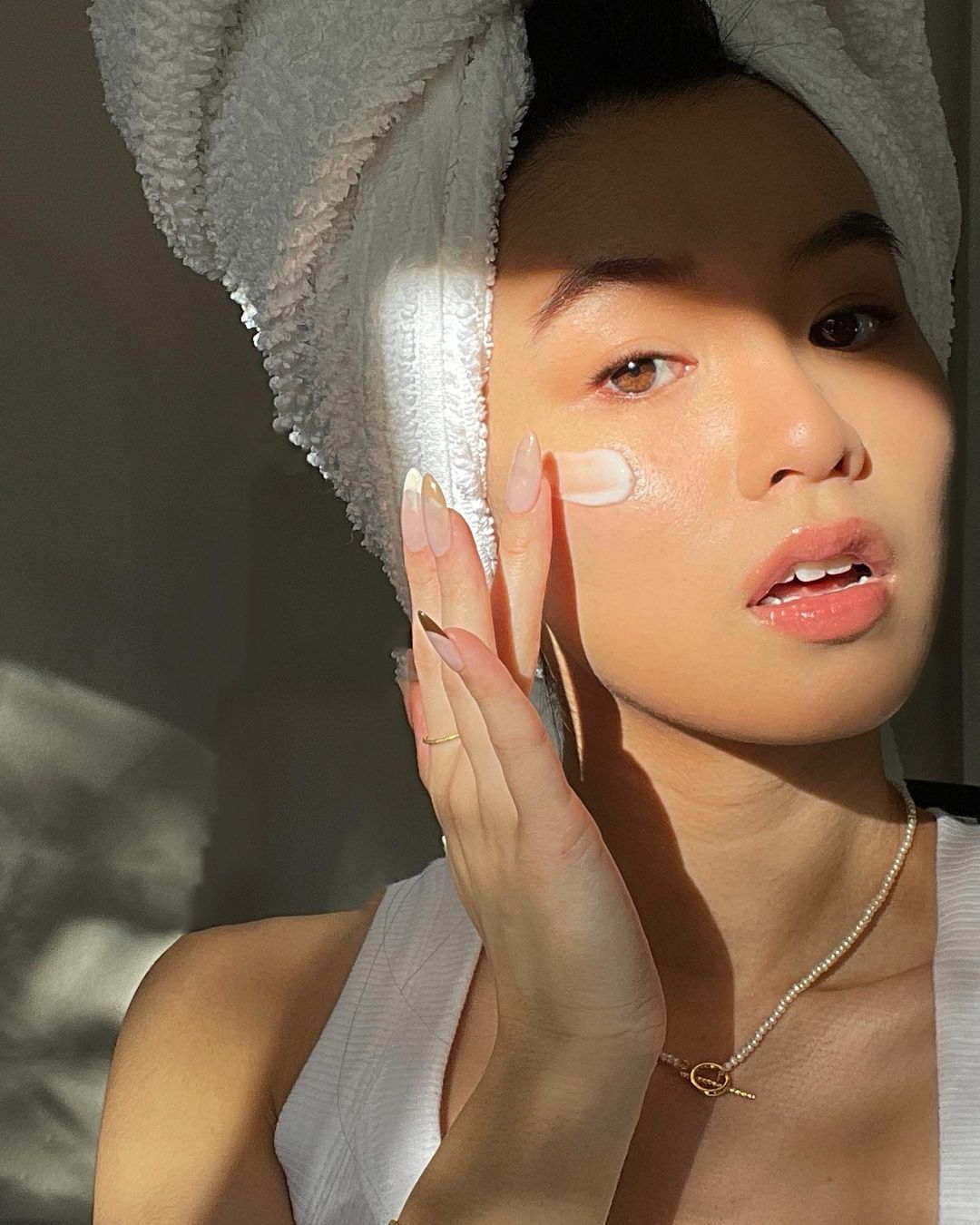 Asian woman applies face cream, wearing a white towel wrap and tank top, in soft light.