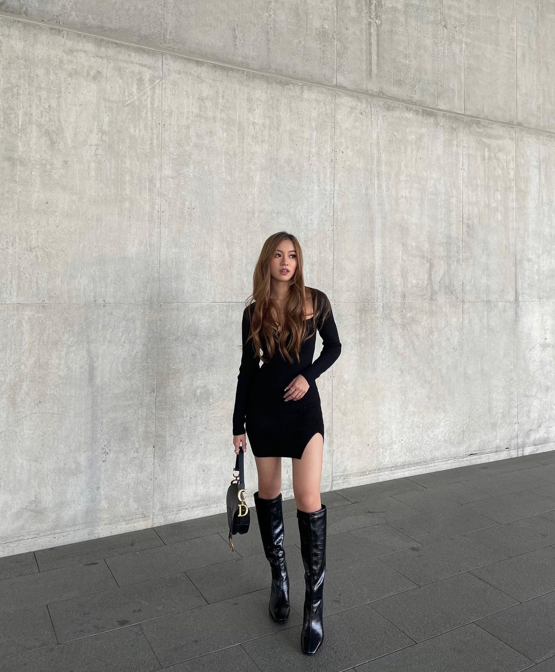 An adult woman stands in a black dress and boots in front of a textured concrete wall, holding a black purse; fashion photography.