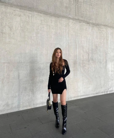 Content creator Ashleigh Huynh in a black dress and boots standing against a concrete wall
