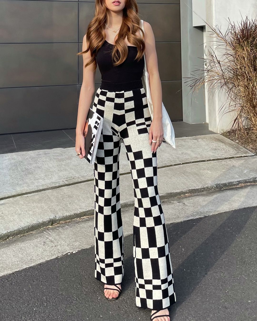 A young woman stands outside in black top and checkerboard pants, holding a book, with a white bag over her shoulder.