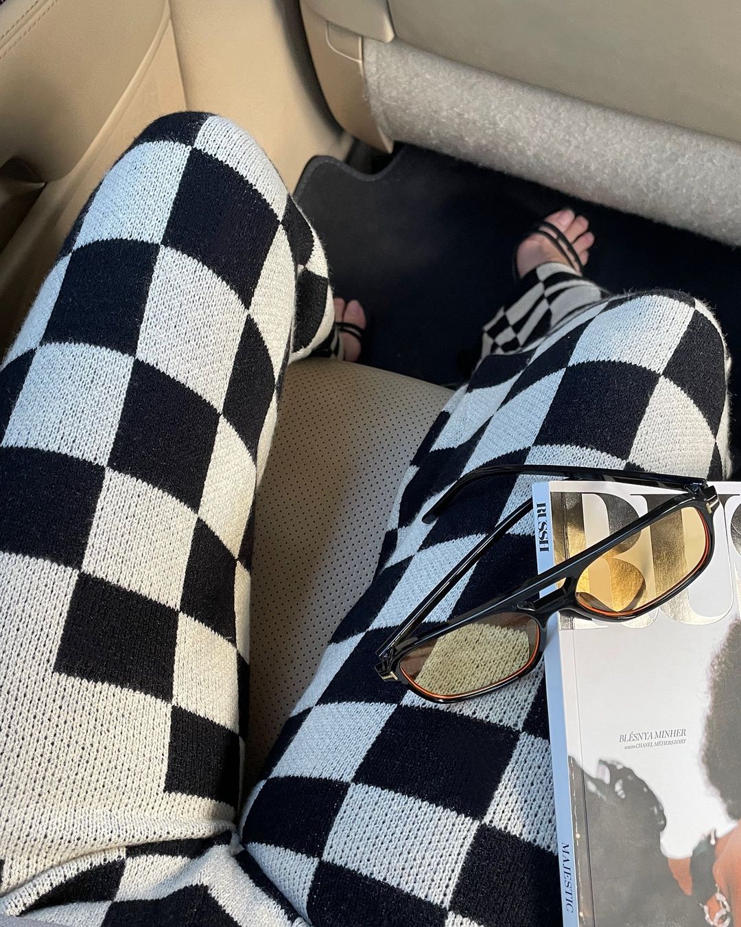 Person seated wearing black and white checkered pants with a magazine and glasses in a car, showcasing a casual fashion style.