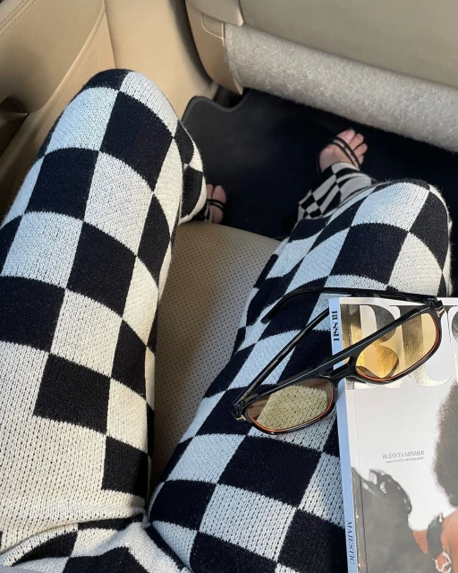 Person seated wearing black and white checkered pants with a magazine and glasses in a car, showcasing a casual fashion style.