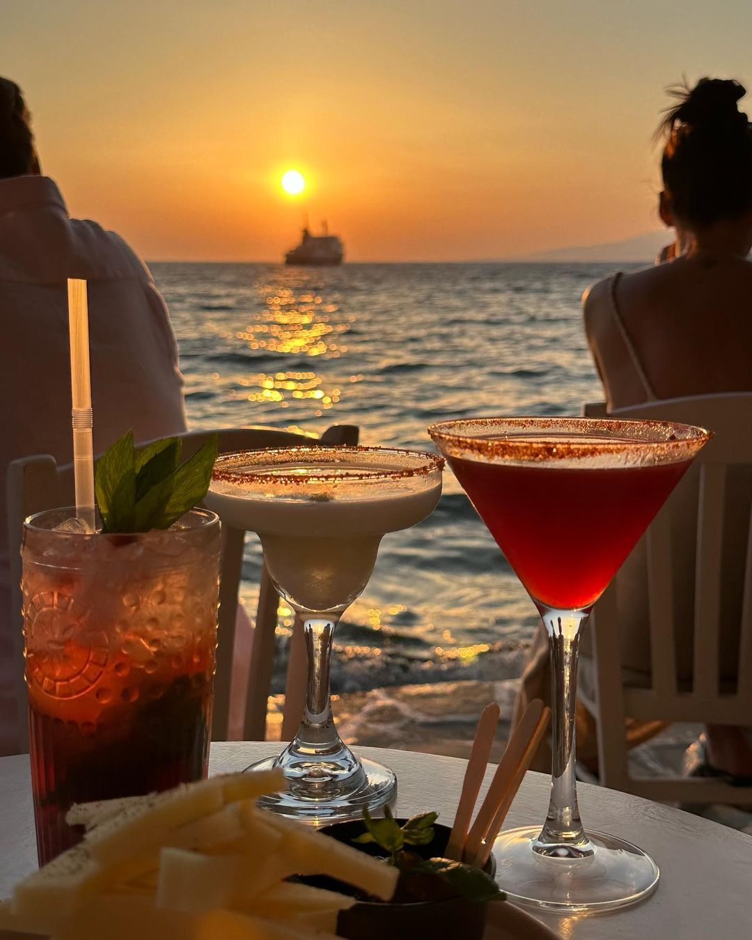 Three cocktails are set on a white table at the beach against a sunset and ocean backdrop with a distant cruise ship on the water.