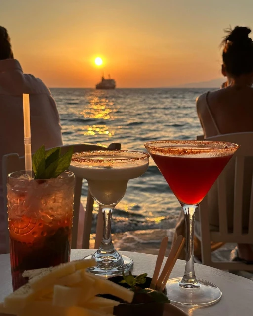 Three cocktails are set on a white table at the beach against a sunset and ocean backdrop with a distant cruise ship on the water.