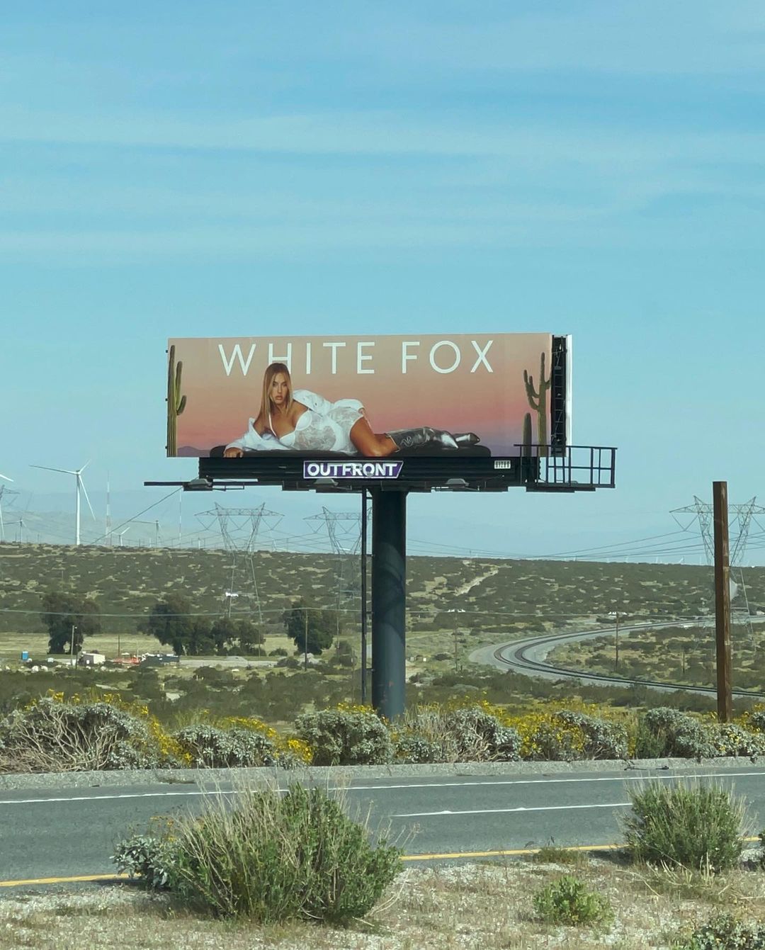 Billboard on a rural highway featuring a blonde model in a white outfit advertising White Fox, under a sunny blue sky landscape.