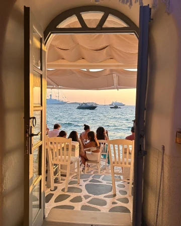People seated at a seaside restaurant with open door framing view of boats on water at sunset, creating a warm, inviting atmosphere.