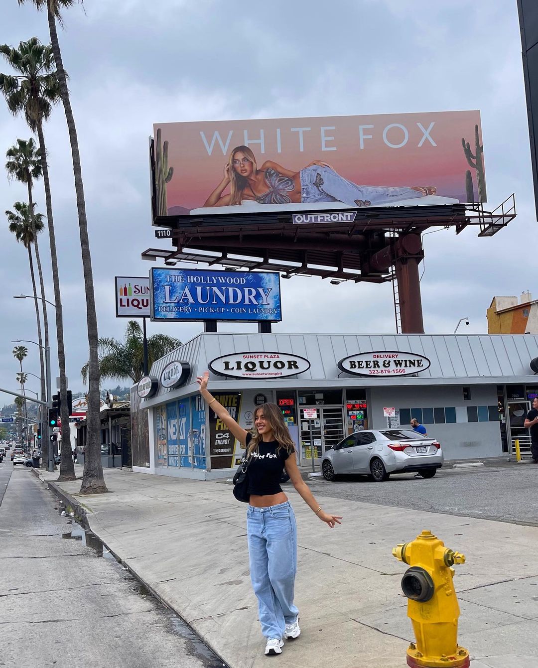 Woman in jeans walks on a sidewalk near palm trees and a billboard for White Fox with urban buildings in the background on a cloudy day.