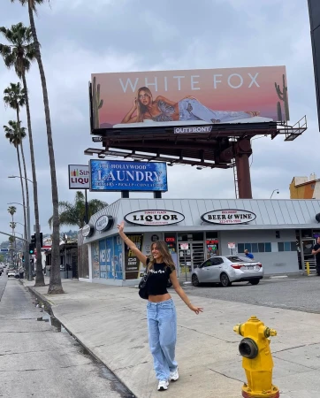 A woman in a black crop top and loose blue jeans waves on a city sidewalk beneath a large billboard.