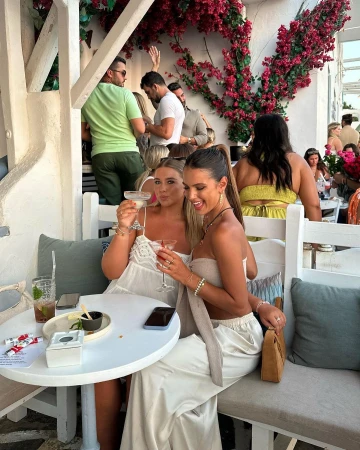 Two women in light-colored outfits hold cocktails while seated at a table at an outdoor restaurant with several people in the background.