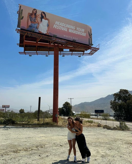 Two young women hugging in front of a White Fox billboard featuring fashion models, with a desert landscape and blue sky backdrop.