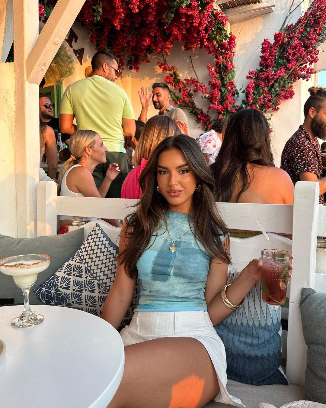 Brunette woman lounges outdoors with cocktail. Red flowers adorn the bright background. People socialize casually.