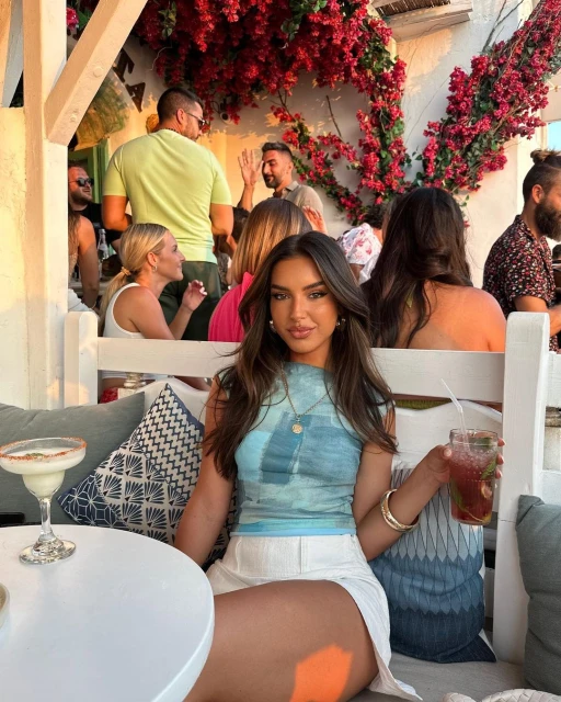 Brunette woman lounges outdoors with cocktail. Red flowers adorn the bright background. People socialize casually.