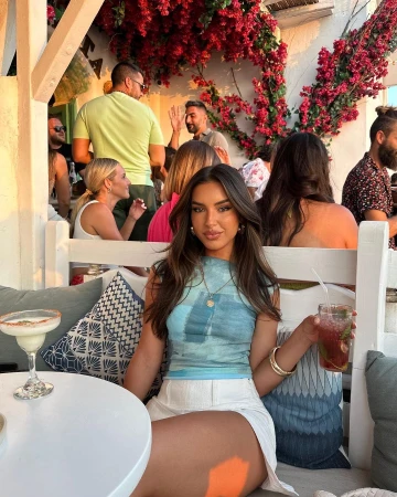 Brunette woman lounges outdoors with cocktail. Red flowers adorn the bright background. People socialize casually.