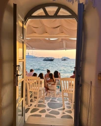 View through an open arched doorway onto a seaside terrace with people seated at tables overlooking the ocean at sunset.