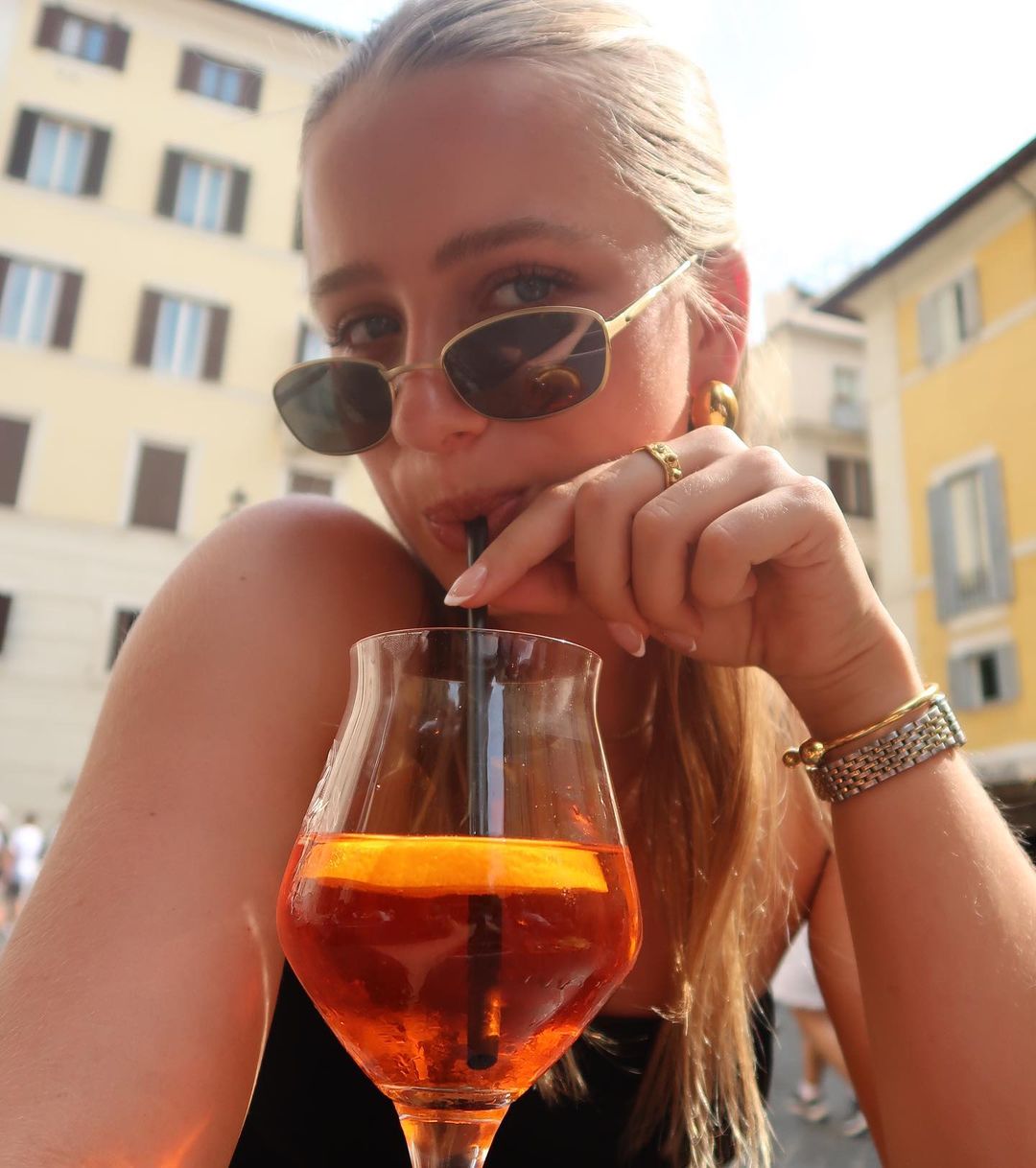 Adult female in sunglasses drinking an orange-colored beverage in a wine glass outside in front of a building on a sunny day.
