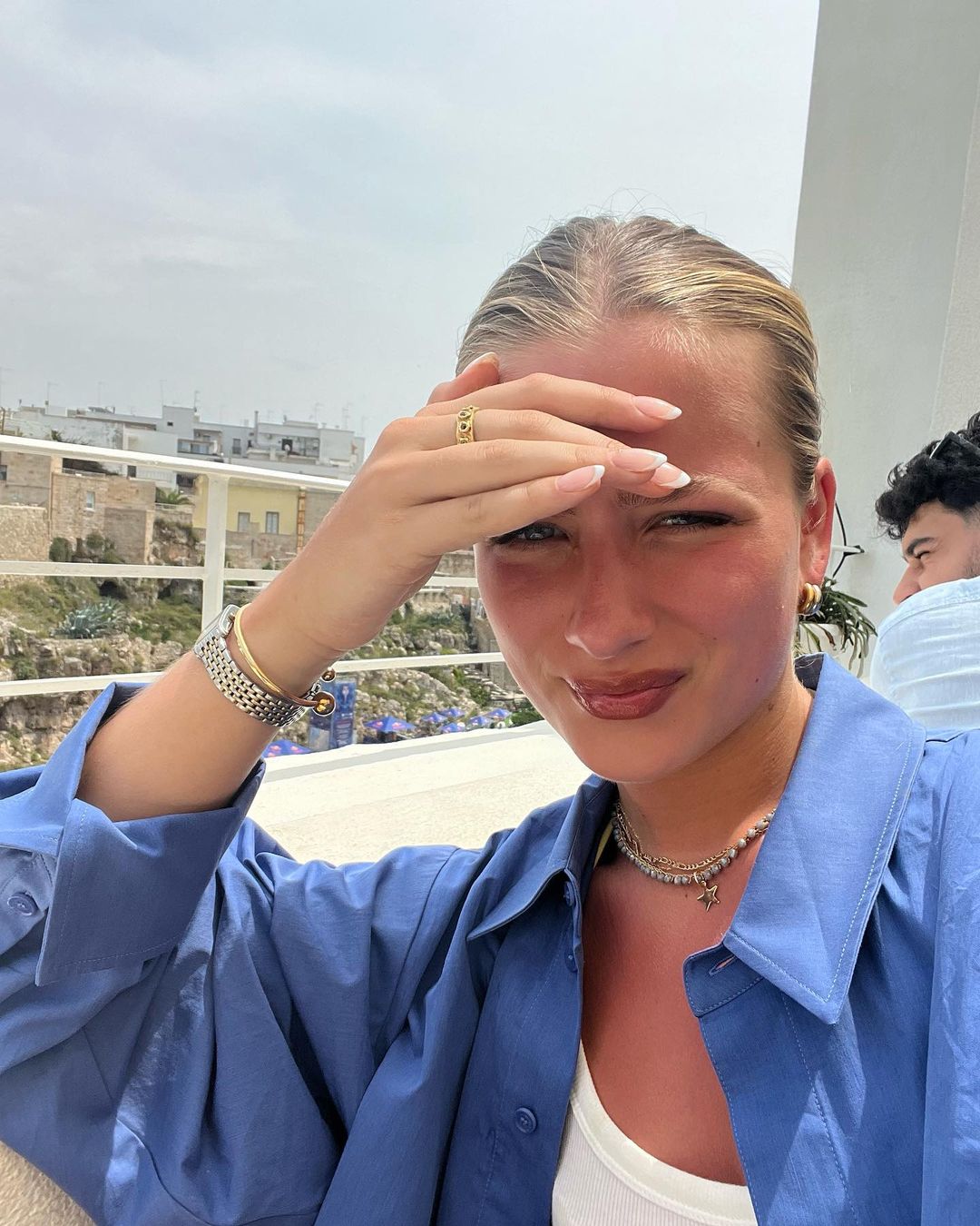 A young fair-skinned woman shades her eyes in an outdoor setting, wearing a blue button-down shirt, with buildings in the background.
