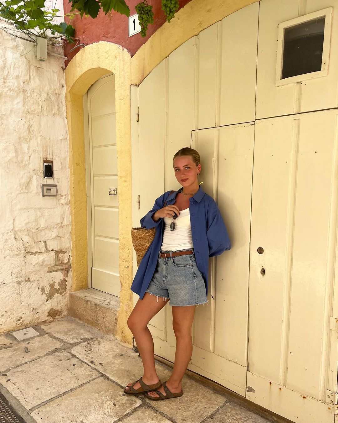 Young woman in blue shirt and jean shorts standing in front of a white door next to a white stone wall with grapes.