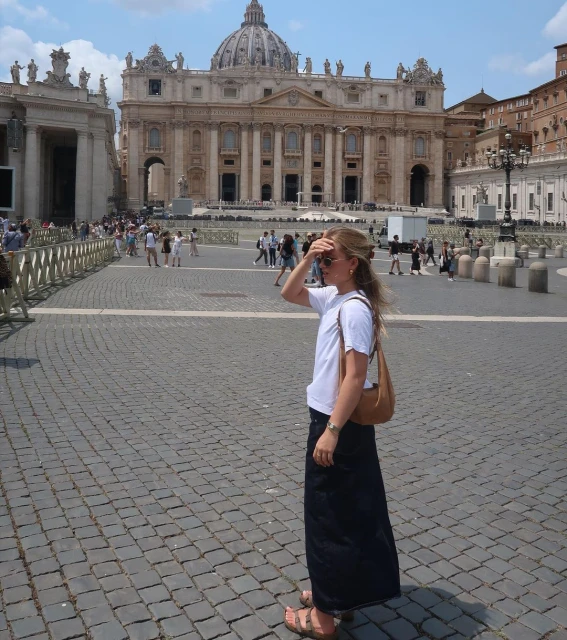 Woman in Piazza Pio XII, Rome, stands before St. Peter's Basilica. Paved square, bright daylight, blue sky, architectural landmark.