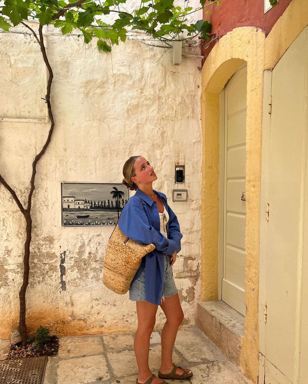 Blond woman in denim shorts and blue shirt looks up next to a yellow door, accessorized with a straw bag in an alley with trees.