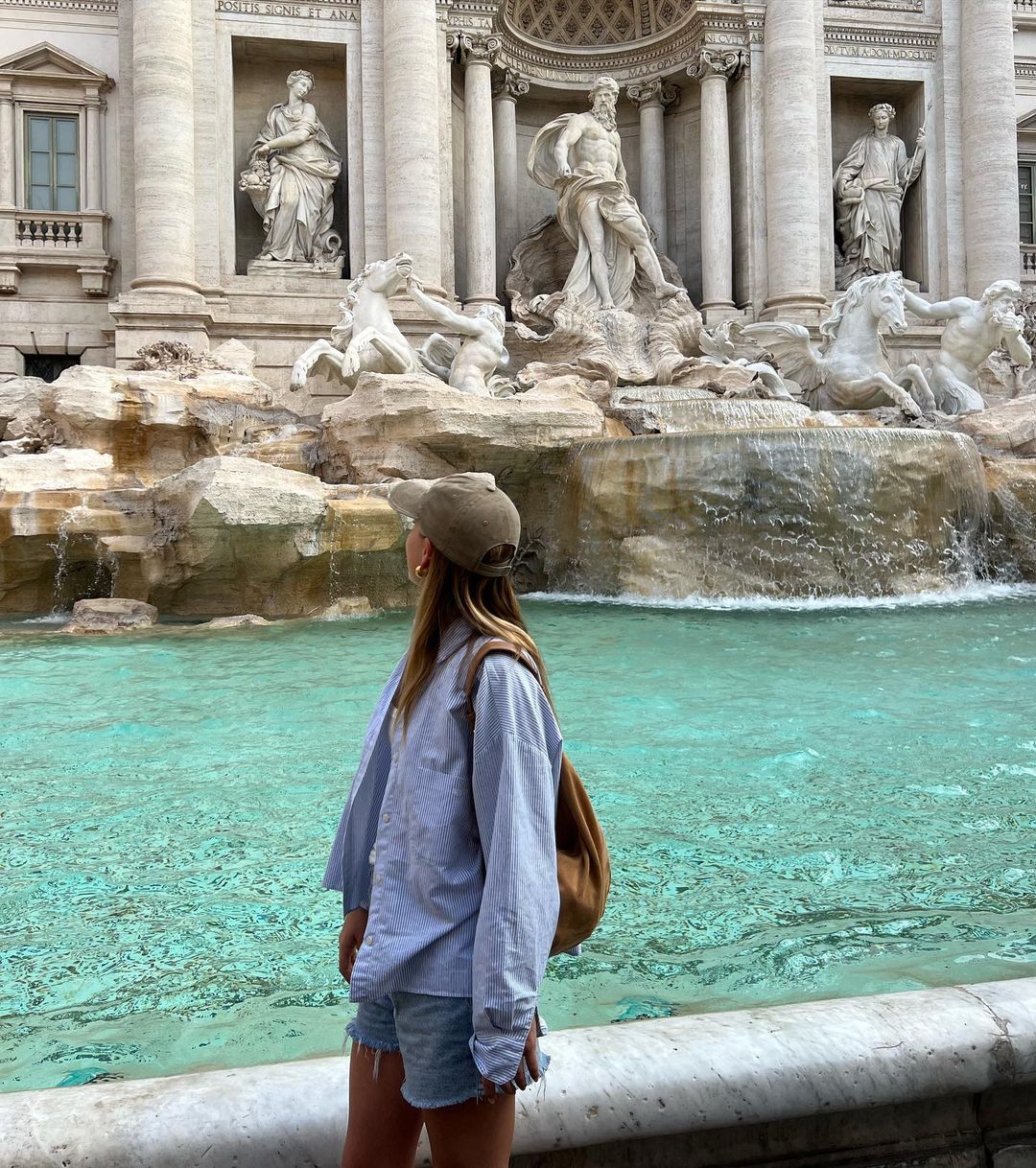 A woman wearing a cap and casual clothing stands at the edge of the Trevi Fountain in Rome, Italy, looking out at the water and sculptures.
