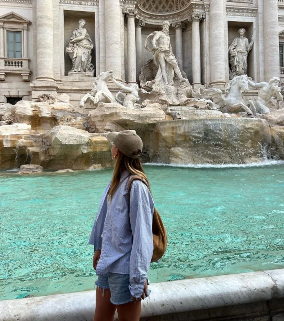 A woman wearing a cap and casual clothing stands at the edge of the Trevi Fountain in Rome, Italy, looking out at the water and sculptures.