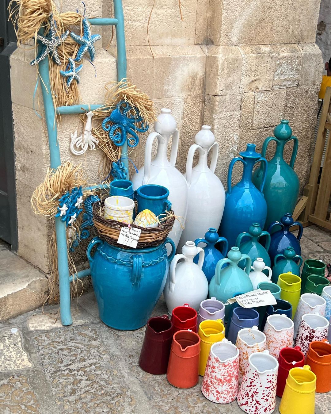 Colorful handcrafted pottery and decorative sea-themed ornaments are displayed outside on a stone surface in a market setting.