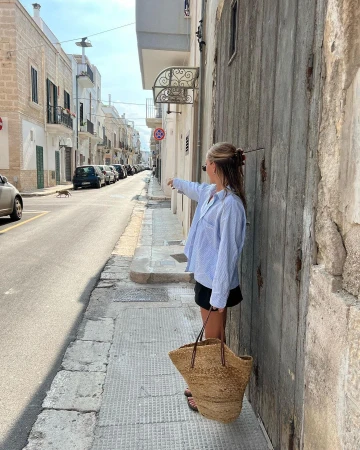 Woman in blue striped shirt and black shorts pointing down a narrow street holding a large straw tote bag