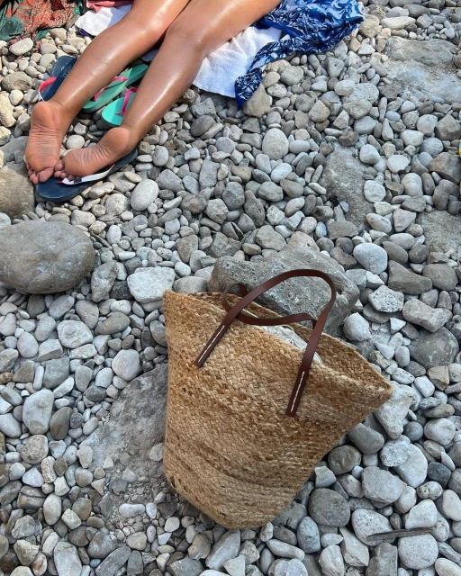 Woman's legs and feet visible while reclining on a rocky beach next to a straw tote bag with leather handles and blue sandals.