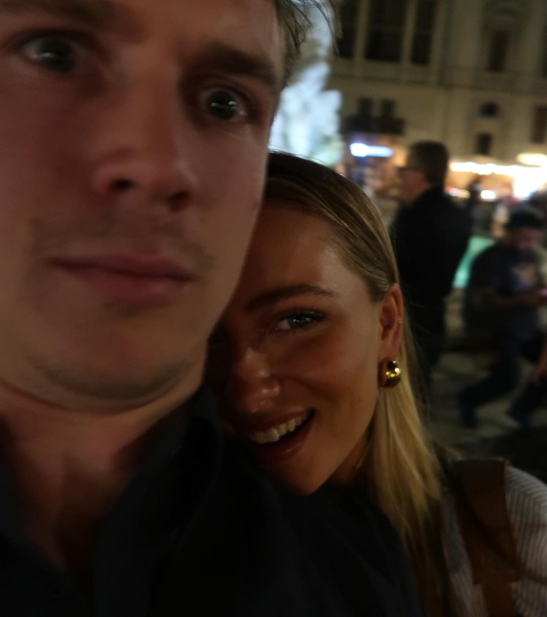 Close-up of a smiling woman with blonde hair next to a man with dark hair, both posing outdoors at night with building lights in background.
