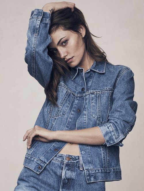 Young woman poses indoors wearing blue denim jacket and pants. She has brown hair and fair skin with a hand on her head.