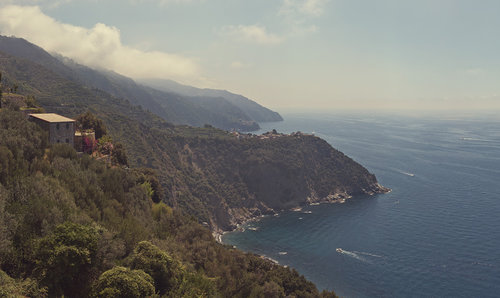 Coastal view features a hillside villa, Mediterranean vegetation, and a scenic ocean backdrop on a clear day with distant boats.