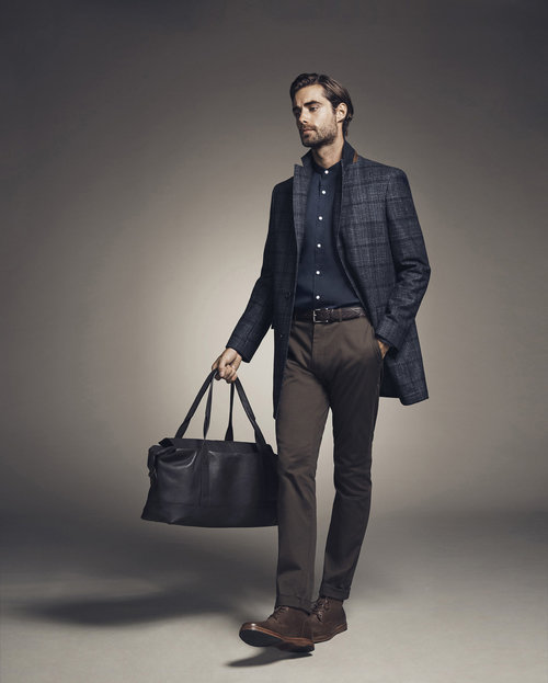 Stylish man walks with a dark leather duffel bag, wearing a patterned overcoat, dark collared shirt, brown pants, and brown leather shoes.