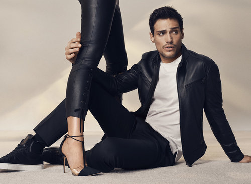 An adult male model poses in a photo studio wearing a black leather jacket with a woman's leg and high-heeled shoe in the foreground.