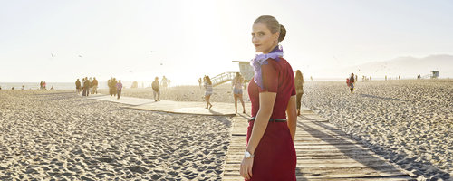 Woman in a maroon dress and purple scarf stands on a wooden boardwalk on a beach with other people in the background.