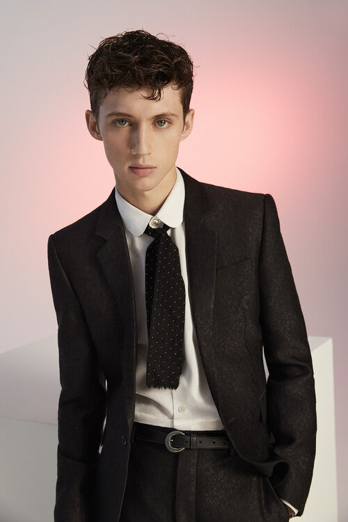 Portrait of adult male with curly brown hair in dark suit with white collared shirt and dark tie in front of pink backdrop.