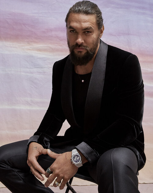 Jason Momoa poses in black velvet suit and watch against a pastel-colored backdrop in a studio portrait for celebrity and entertainment.