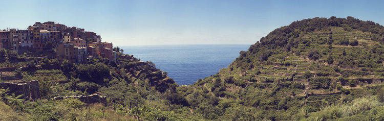 Panoramic view of a coastal village perched on a hillside overlooking the blue sea, surrounded by lush green vegetation and clear blue sky.