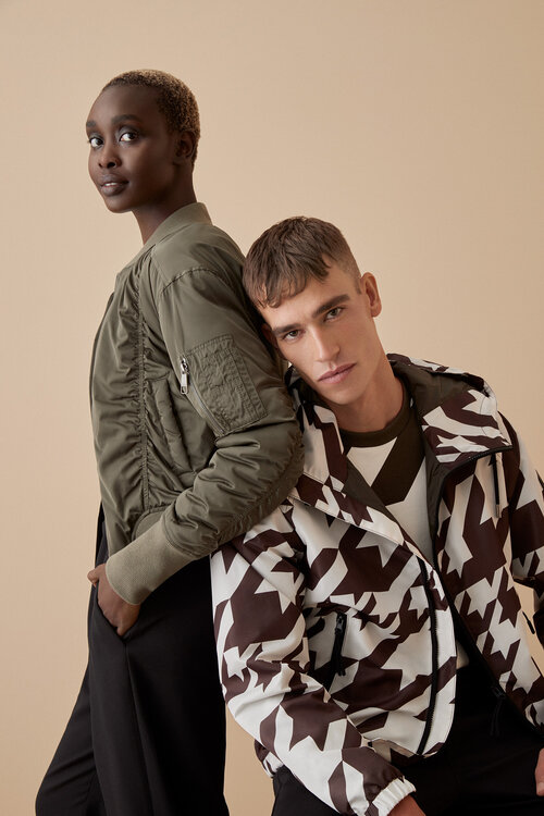 Two fashion models, one Black woman in an olive bomber jacket and one White man in a patterned jacket, pose in front of a tan background.