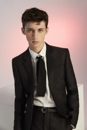 Portrait of a young man wearing a dark suit, white button-down shirt, and a black patterned tie against a gradient backdrop.