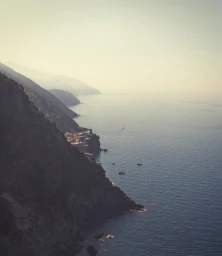 Aerial view of rugged coastal cliffs meeting the ocean with small boats floating on the water below under a hazy sky.