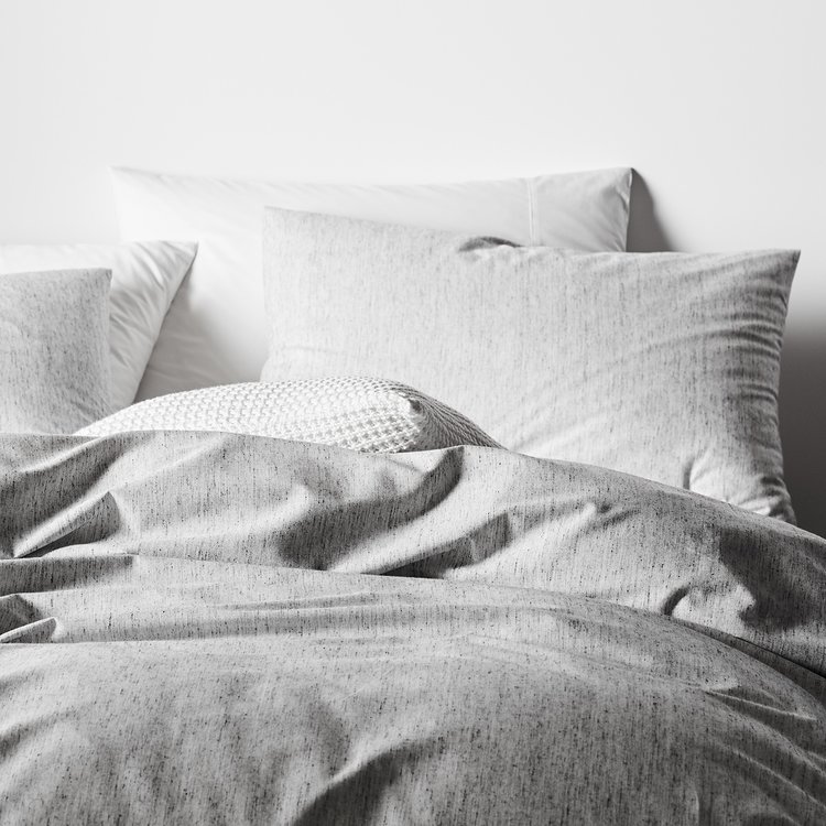 Close-up of a comfortable bed with soft, textured gray and white bedding and pillows against a plain white wall in a bedroom setting.