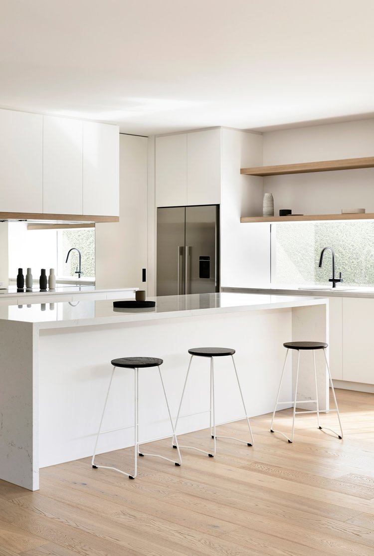 Bright kitchen interior with island, three minimalist stools, white cabinets, stainless steel refrigerator, light hardwood floor.