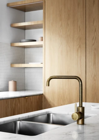 Modern kitchen interior featuring a brass faucet, stainless steel sink, marble countertop, wood cabinets, and white tiled backsplash.