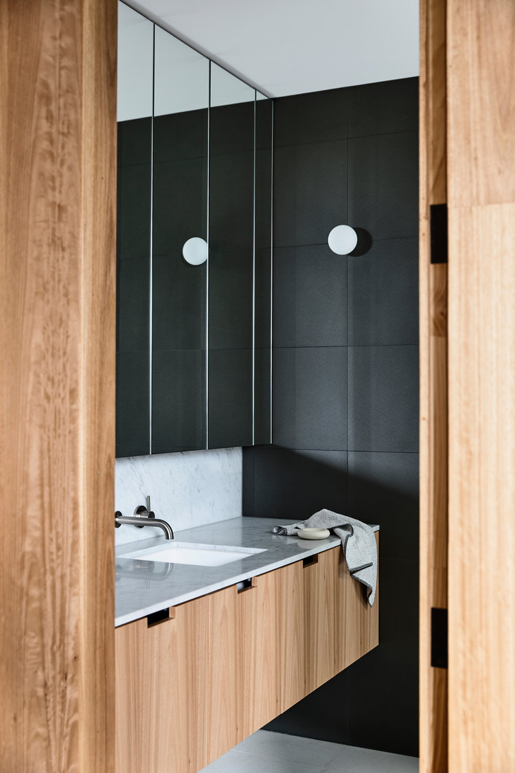 Contemporary bathroom interior featuring a wood vanity with a marble countertop, a white sink, and a black tiled wall with round lights.