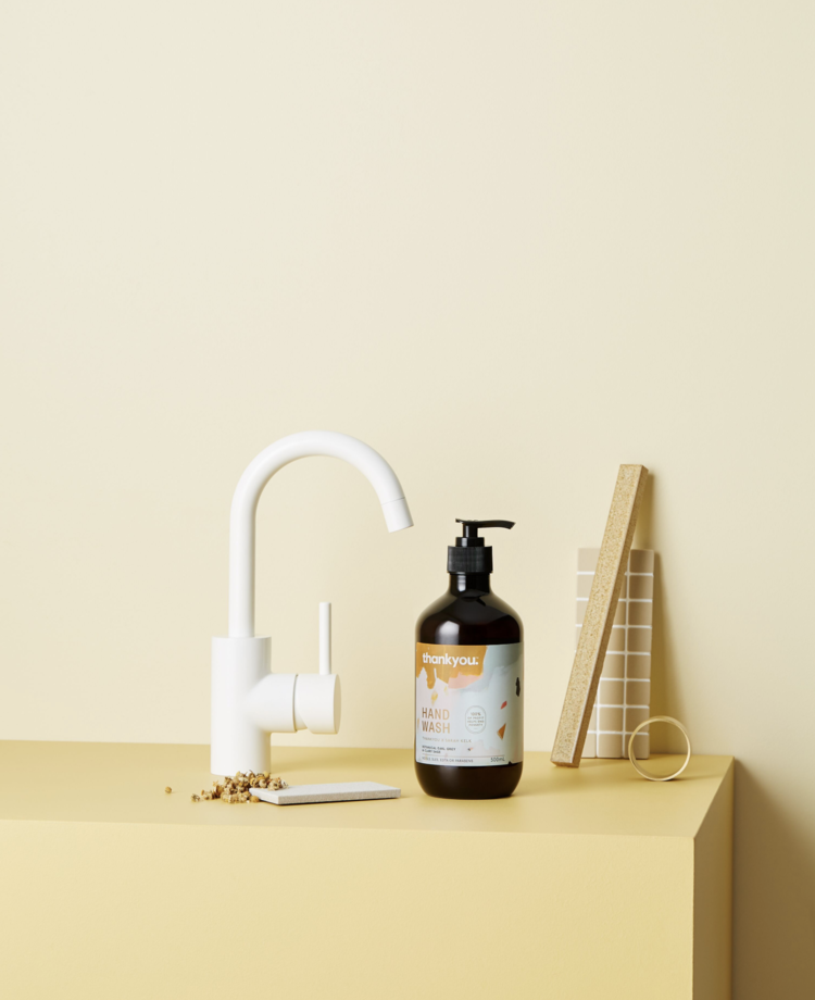 Still life shot of a white faucet and brown hand soap bottle, balanced with a wooden block, ceramic tile, and golden ring on a yellow surface.