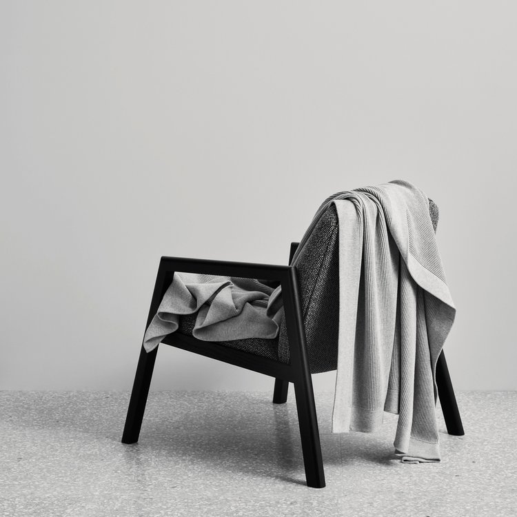 Black wood chair draped with light grey woven blankets in a neutral interior with speckled floor, minimalist furniture photography.