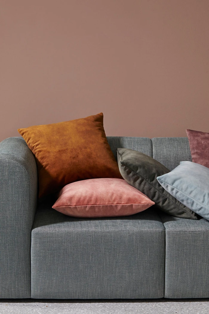Close-up of a grey upholstered sofa styled with colorful velvet cushions in rust, pink, olive, and light blue against a mauve wall