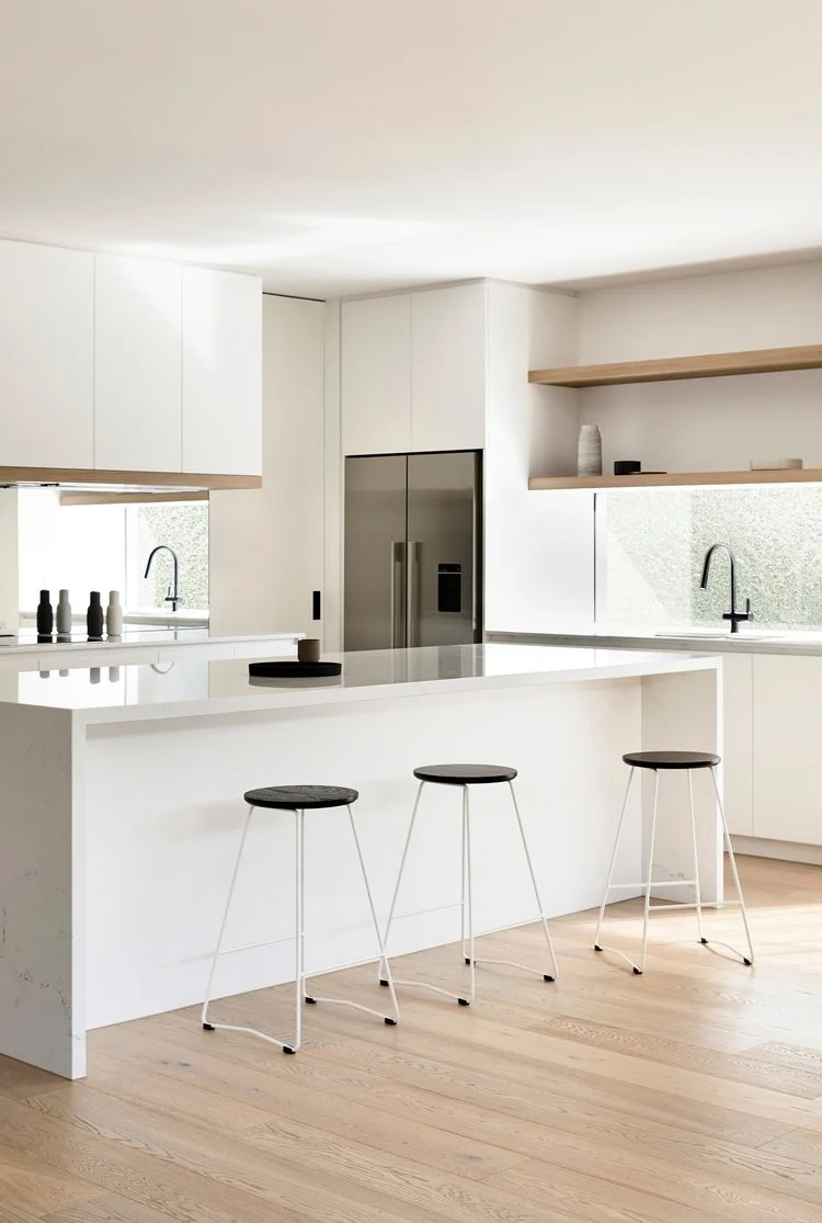 Modern minimalist kitchen interior with white cabinets, a large island bench, wooden open shelving, and three black and white bar stools.